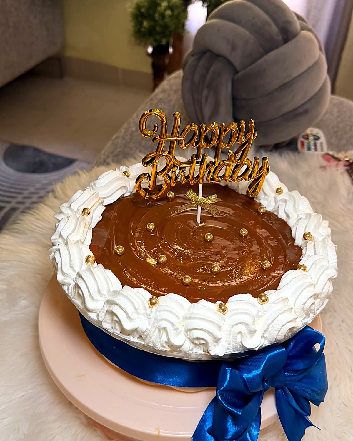 A round cake with its top covered in dulce de leche and gold pearls, a peaked white border, and a blue ribbon at the base.