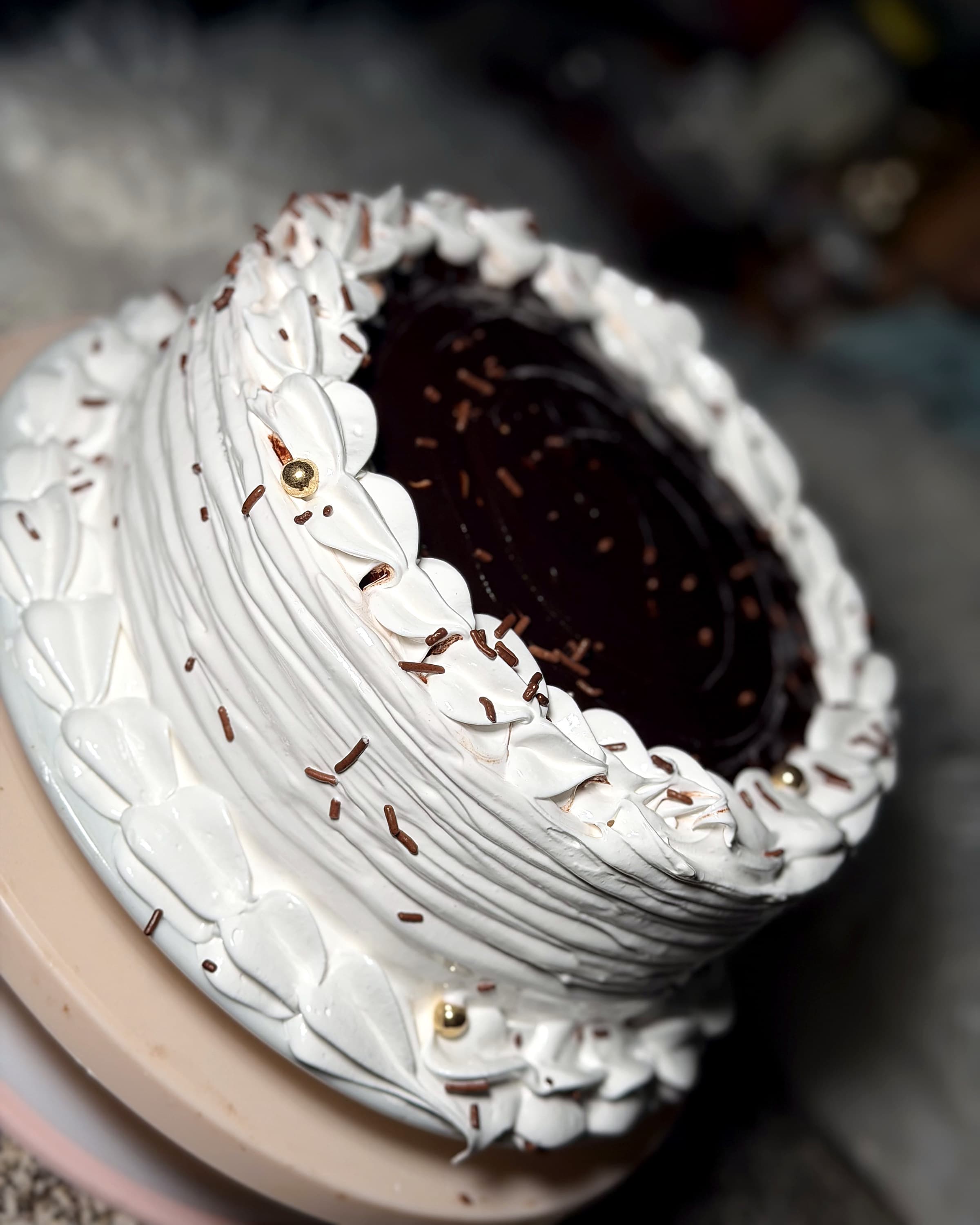 A cake with a glossy chocolate ganache top scattered with chocolate sprinkles, inside a piped white meringue border of petals, pleated sides, and a few gold pearls.