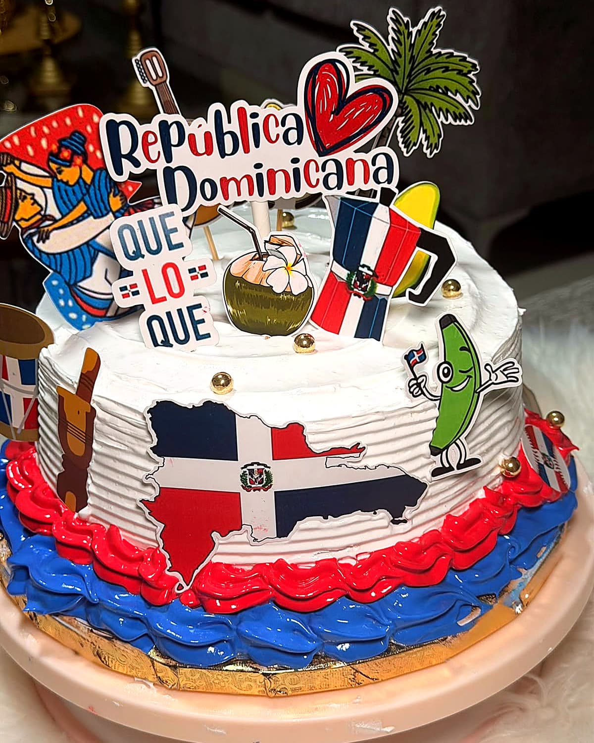 A white cake carrying the Dominican map and flag on the front, red and blue piping around the base, and a topper reading “Que lo que”.