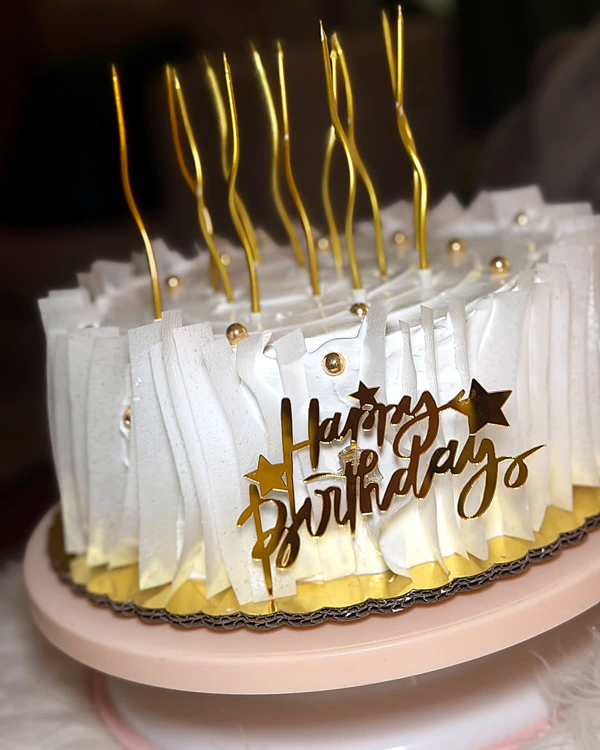 A white cake with pleated frosting, a row of wavy gold candles, and a gold “Happy Birthday” topper.