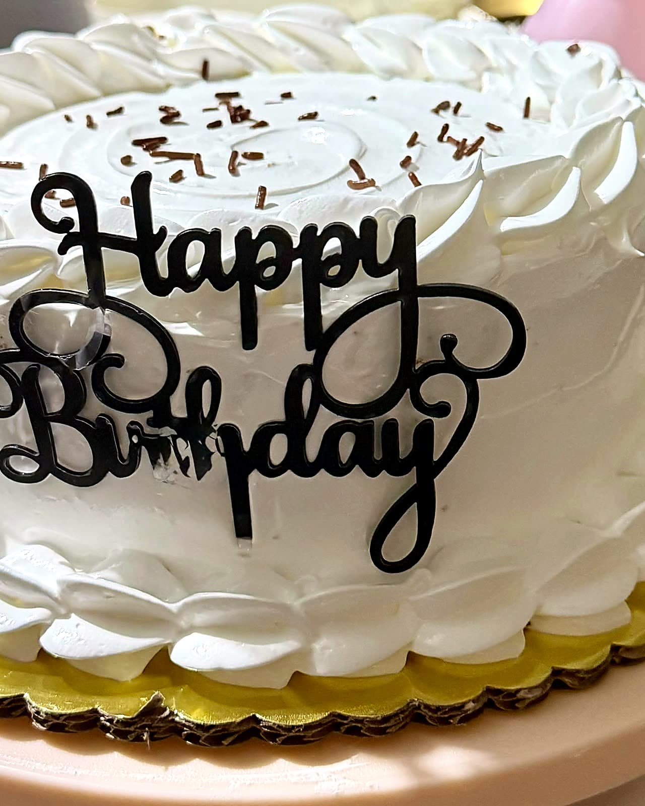 A white cake scattered with chocolate shavings, with a black “Happy Birthday” topper.