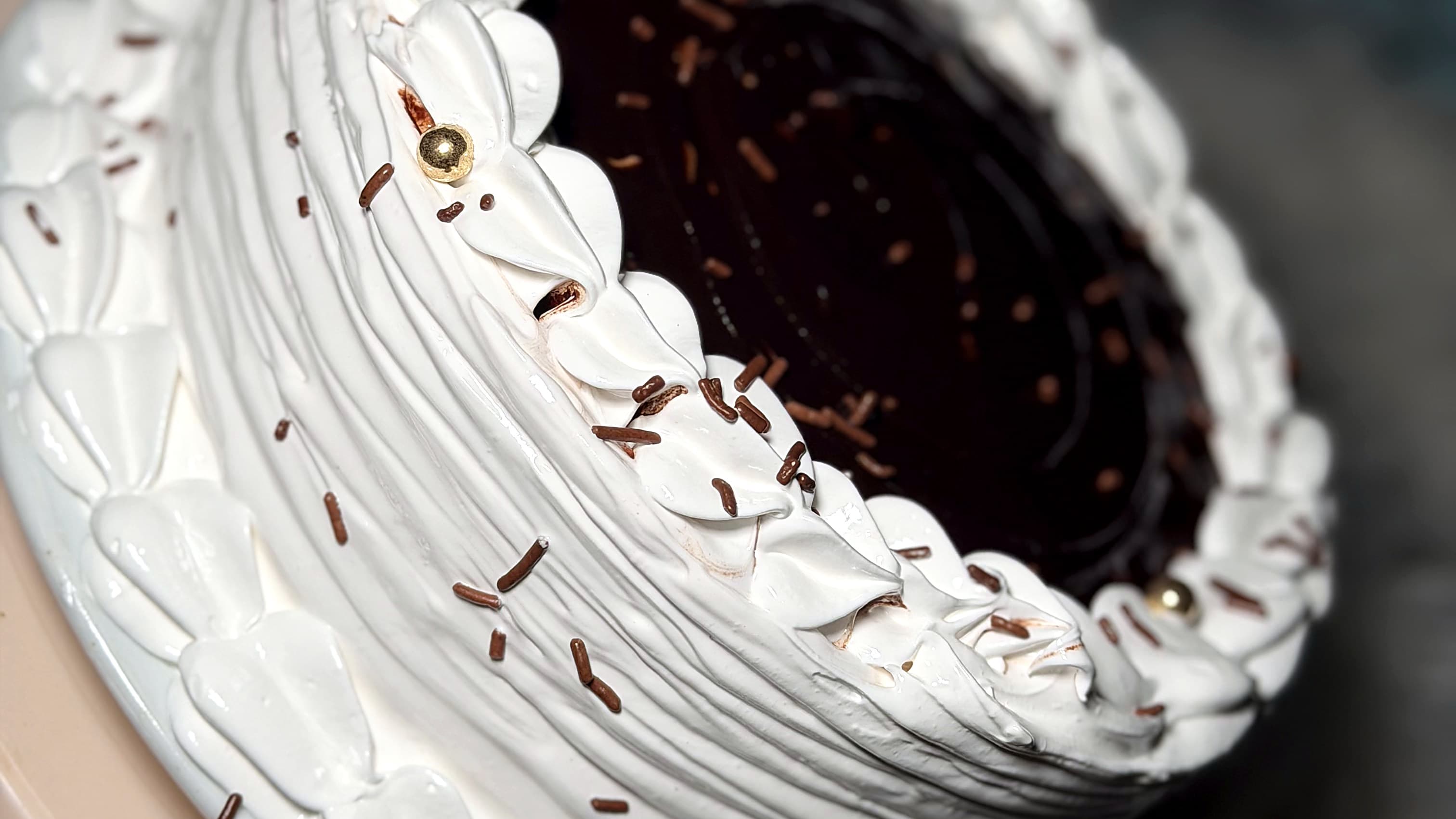 Bizcocho con el tope de ganache de chocolate brillante y fideos de chocolate, dentro de un borde de merengue blanco en pétalos, los lados en pliegues y unas perlas doradas.